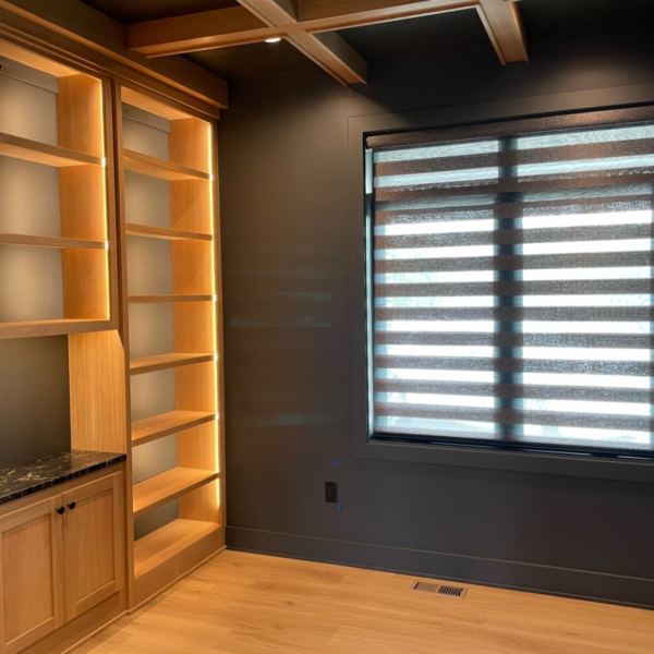 Custom home office with illuminated built-in shelving, coffered ceiling, and modern window treatments.